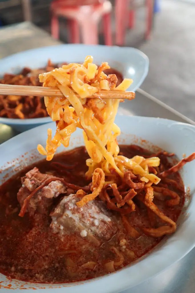คีบเส้นข้าวซอยขึ้นจากชามน้ำแกงข้นสีแดงของร้านข้าวซอยแม่มณี เชียงใหม่