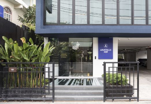 ทางเข้าหลัก Apartelle Jatujak Hotel ป้ายสีน้ำเงิน ล้อมรอบด้วยต้นไม้สีเขียวสด