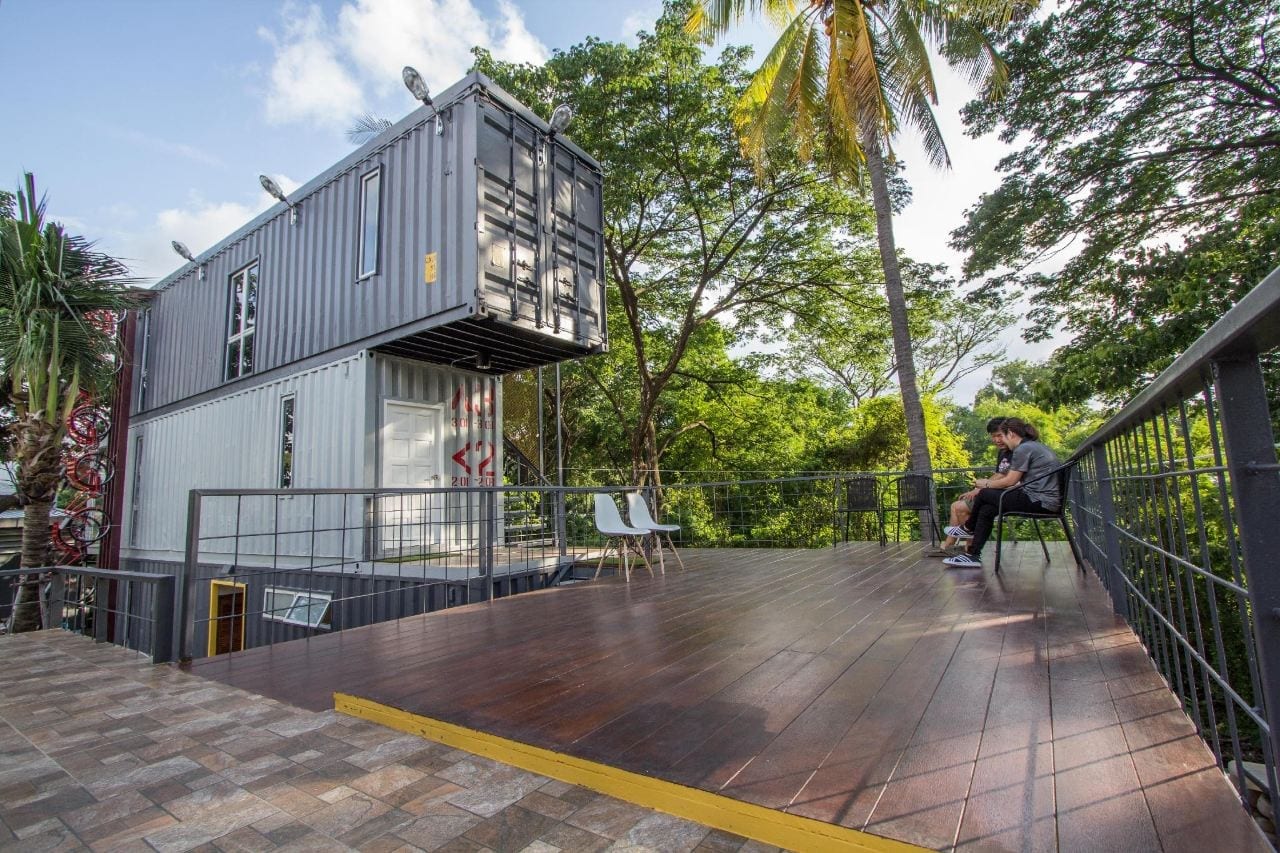 ลานระเบียงไม้และอาคารตู้คอนเทนเนอร์สามชั้นของ Ducati Bike Box Hostel ท่ามกลางต้นไม้เขียว