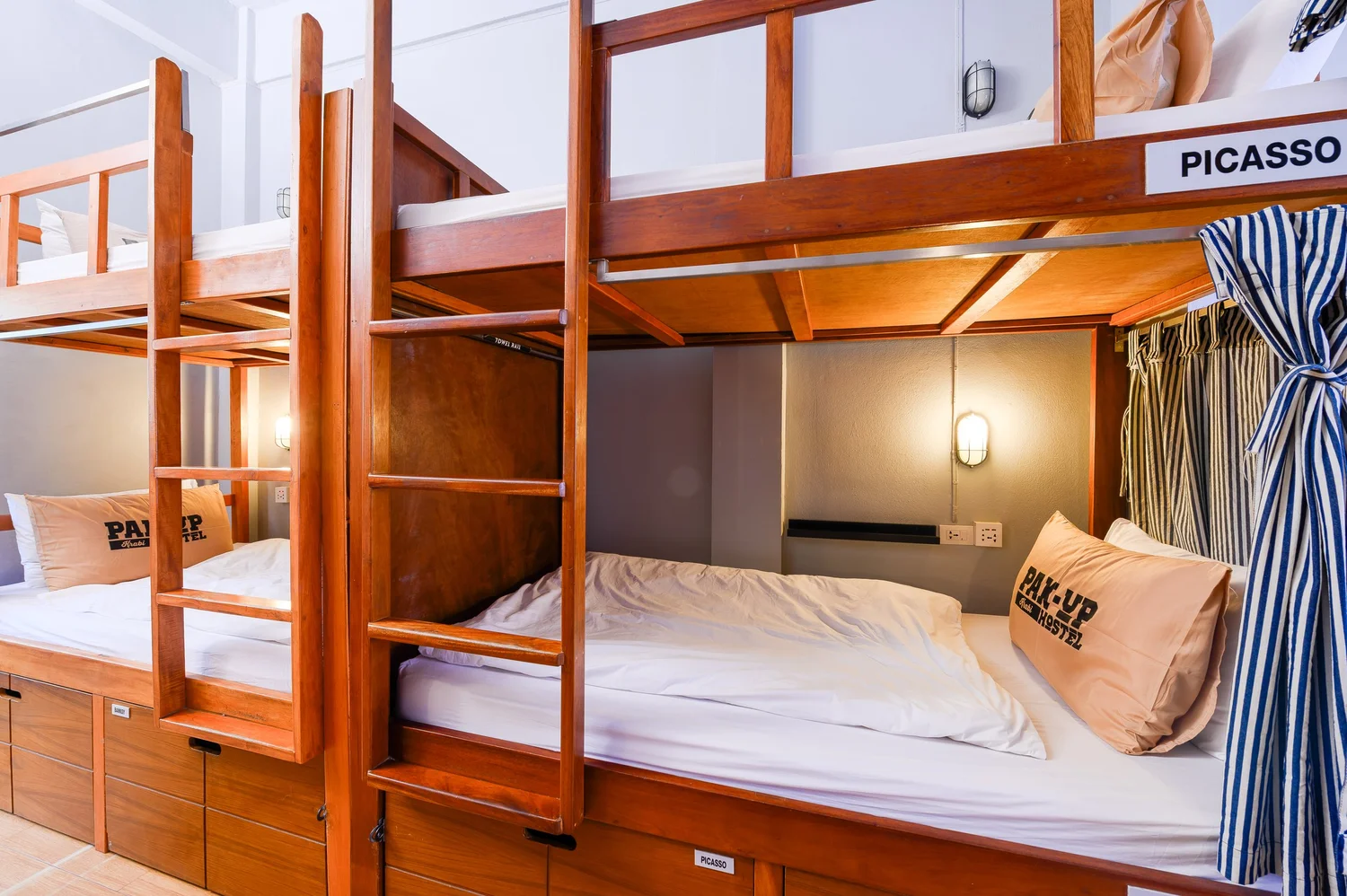ดอร์ม Picasso ของ Pak-Up Hostel เตียงบังก์ไม้มีไฟอ่านหนังสือ ม่านลายทาง และล็อกเกอร์ใต้เตียง
