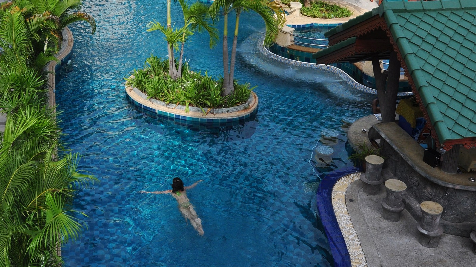 สระว่ายน้ำสไตล์ลากูนของ Baan Karonburi Resort มองจากมุมสูง มีเกาะต้นปาล์มกลางสระและสวิมอัพบาร์หลังคาไทย