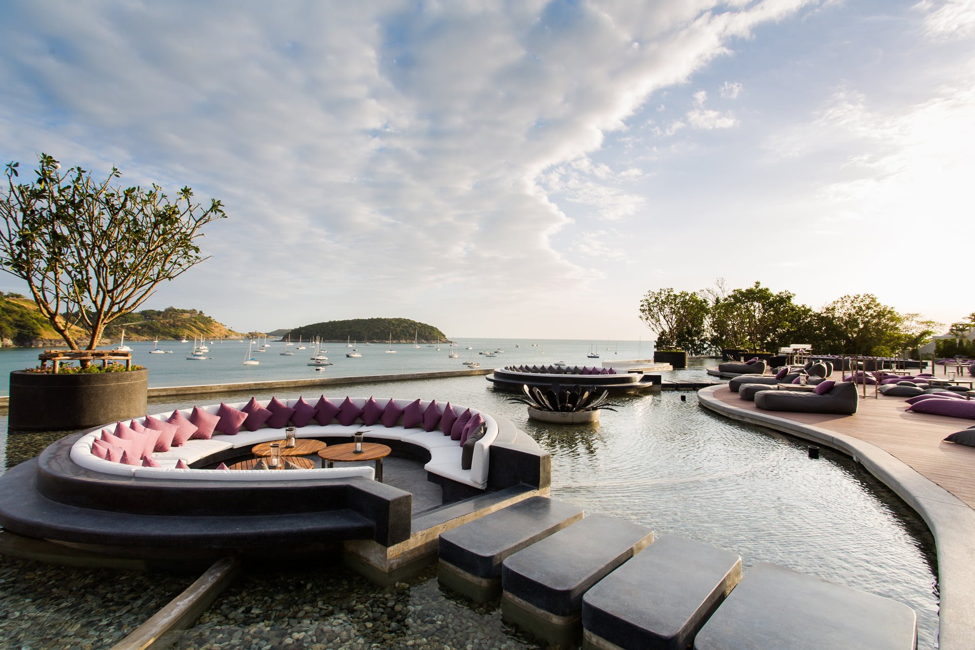 Reflections rooftop bar at The Nai Harn at dusk, curved sofas beside a reflecting pool with yachts in Nai Harn Bay beyond