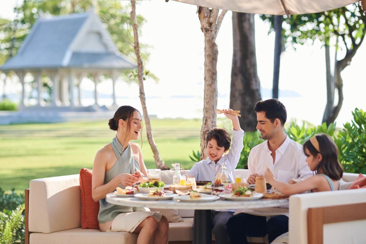 ครอบครัวนั่งกินมื้อกลางวันบนสนามหญ้าริมทะเลของ Centara Reserve Samui โดยมีศาลาทรงคอโลเนียลสีขาวเป็นฉากหลัง