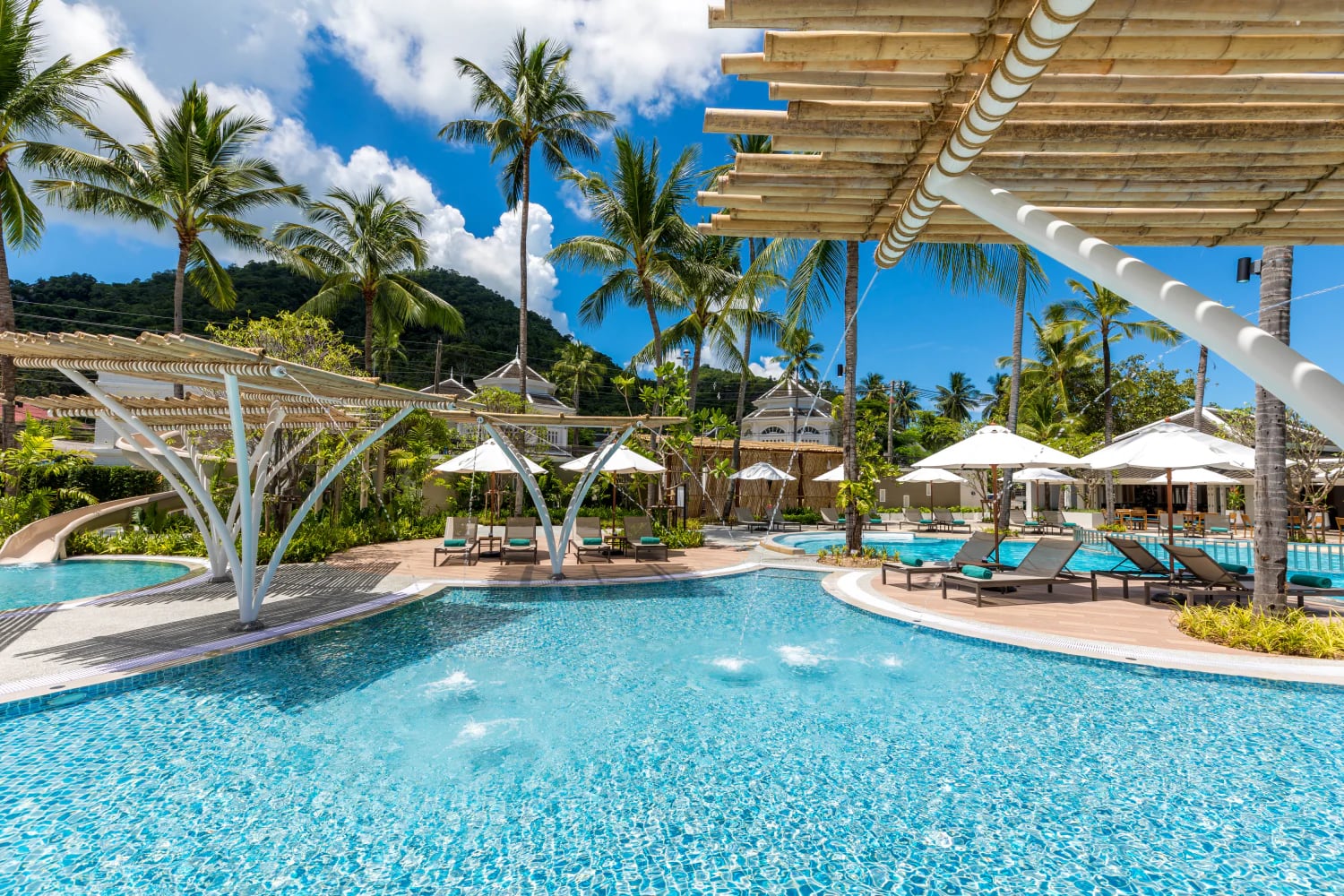The resort-side pool at Outrigger Koh Samui with a children's waterslide and sun shades among the coconut palms, hills behind